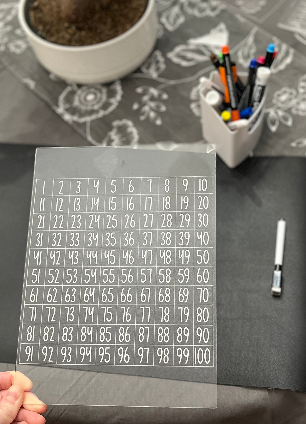 Clear acrylic sheet with a 1 to 100 numbered grid held above a dark surface with markers and a plant in background