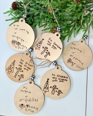 Six round wooden Christmas ornaments with children's drawings and handwritten messages, hanging with twine and plaid beads on pine branches.