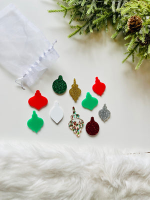 Assortment of colorful glitter and solid acrylic ornament shapes arranged near white pouch and festive greenery with pinecone