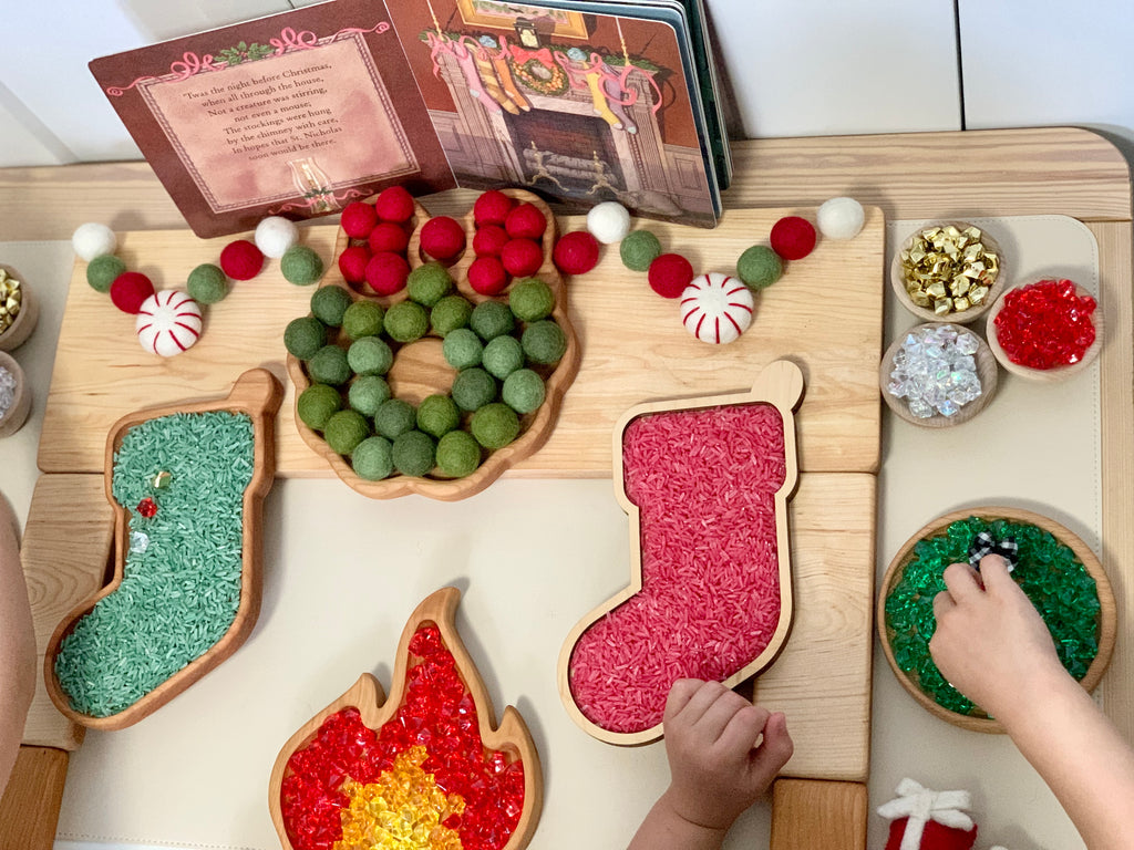 Laser cut wooden stocking-shaped sensory tray with colored rice, felt balls, and festive craft materials on light wood table