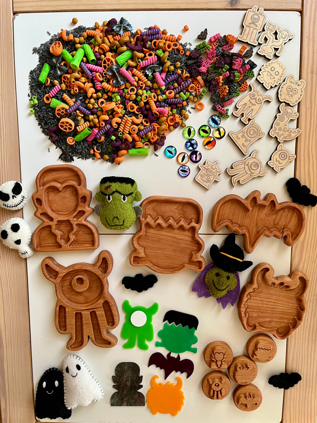 Halloween-themed wooden trays, tokens, colorful pasta shapes, monster eyes, and felt monster figures on white board and wooden table
