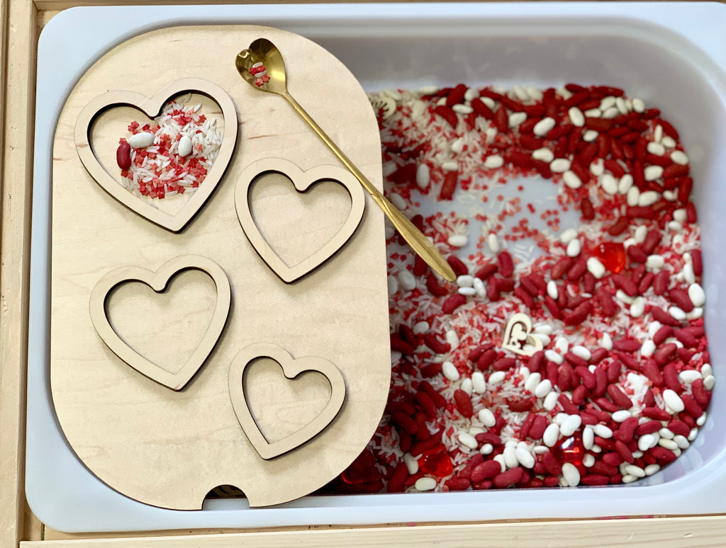 Wooden tray with four heart-shaped cutouts over a bin filled with red and white rice and a gold spoon with a heart-shaped handle