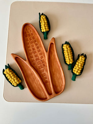 Wooden tray carved into an ear of corn shape with four small felt corn decorations on a beige surface