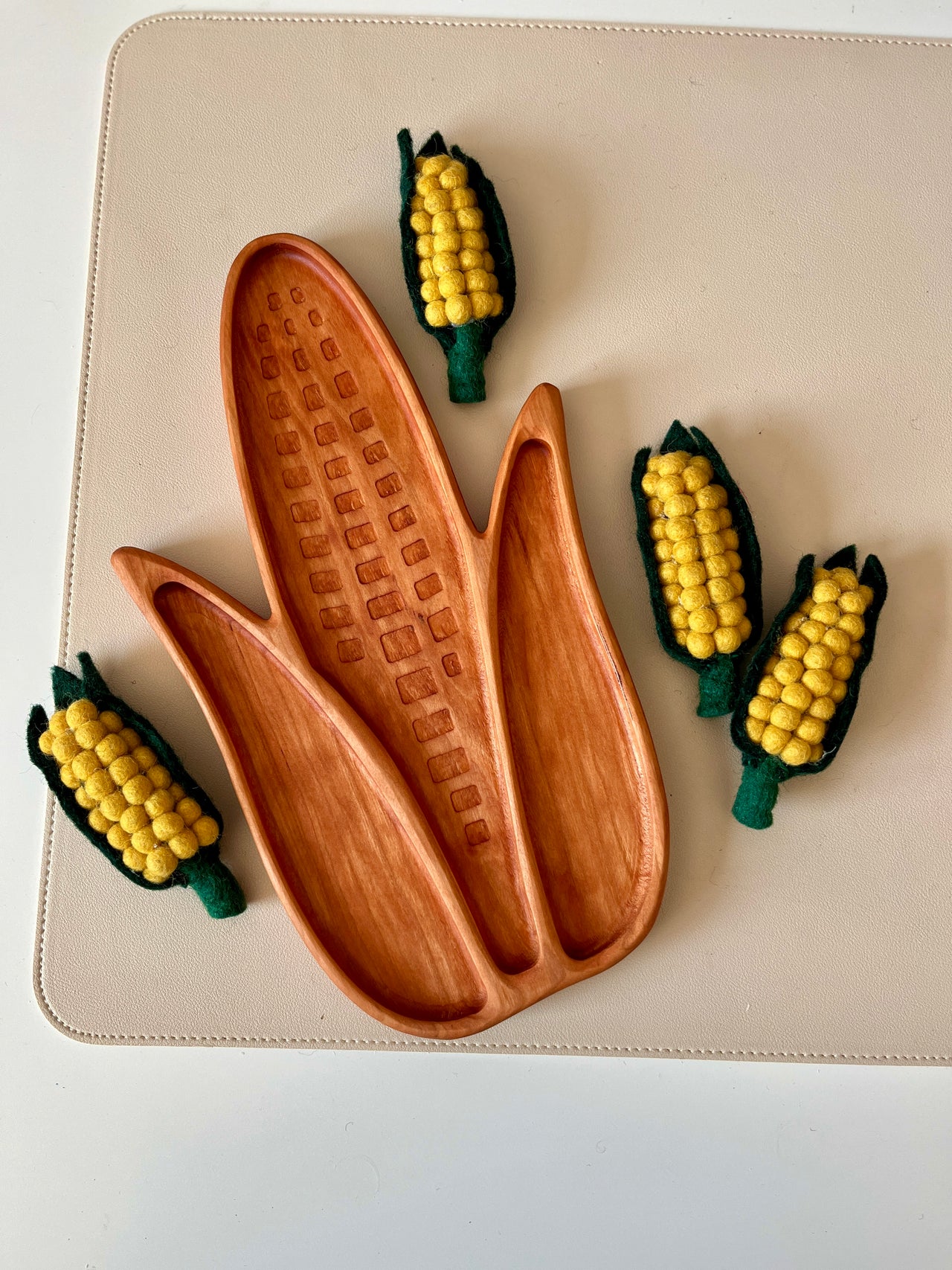 Wooden tray carved into an ear of corn shape with four small felt corn decorations on a beige surface