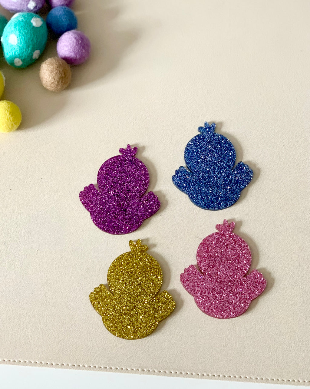 Four glittery chick-shaped cutouts in purple, blue, gold, and pink on a beige surface near colorful felt balls