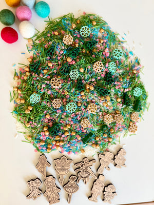 Colorful sensory mix with green pine needles, wooden snowflake shapes, gold beads, and felt balls on white surface with wooden figurines nearby
