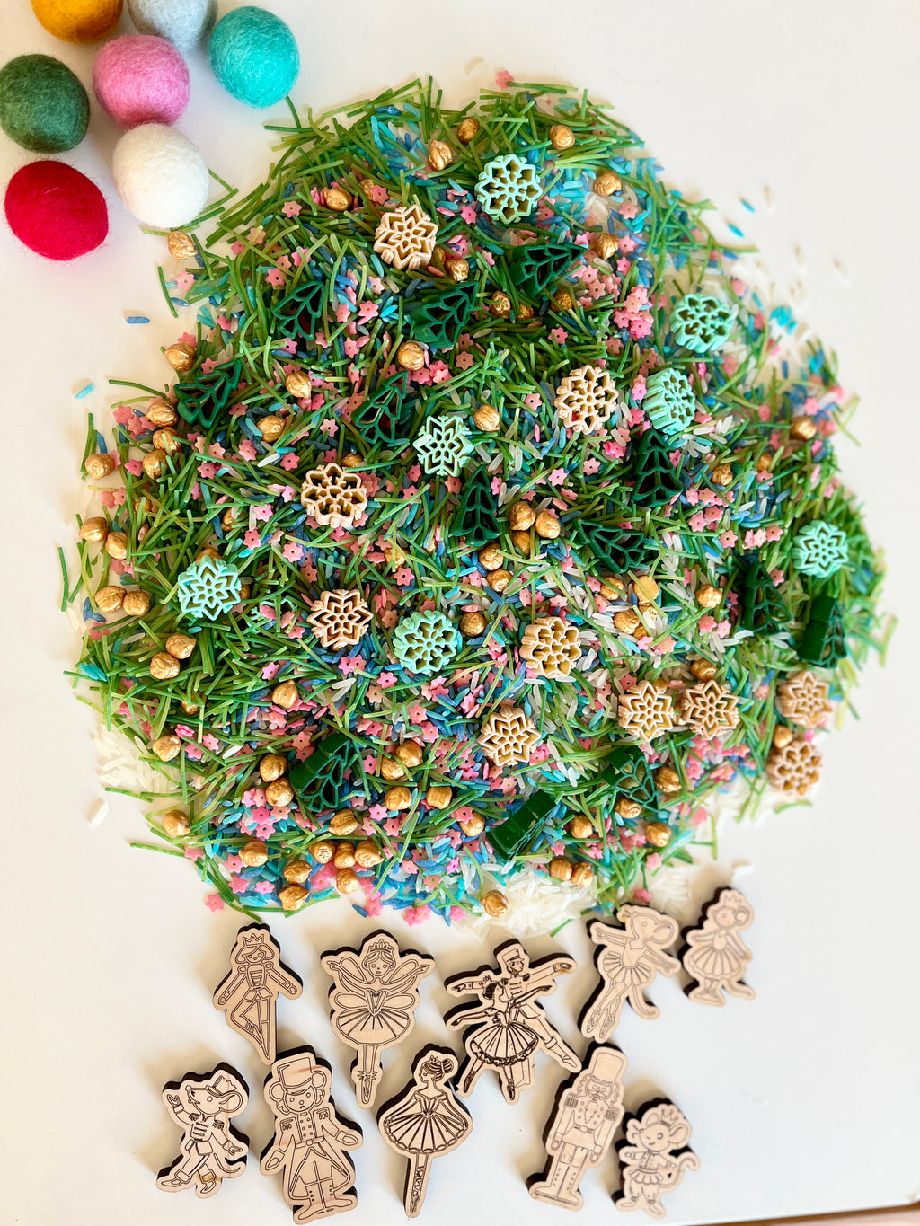 Colorful sensory mix with green pine needles, wooden snowflake shapes, gold beads, and felt balls on white surface with wooden figurines nearby