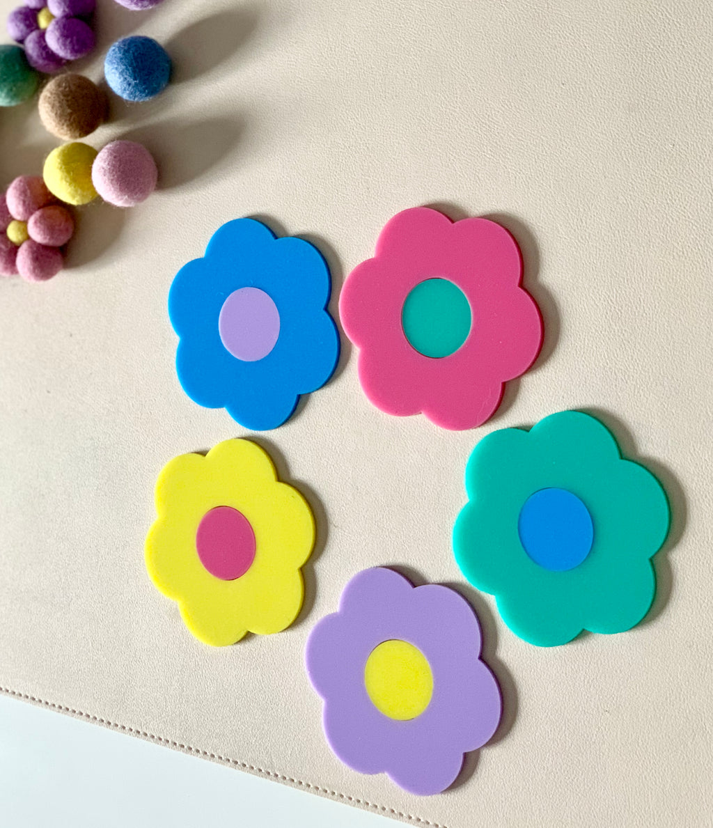Five colorful flower-shaped cutouts in blue, pink, yellow, teal, and purple on a beige surface near felt balls