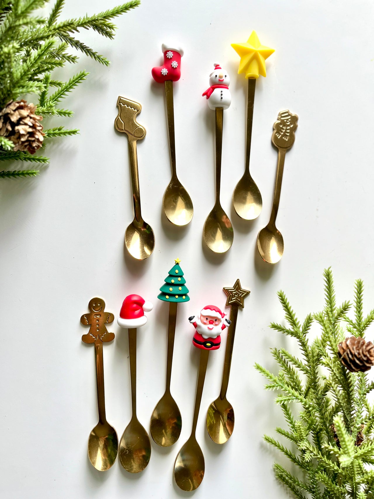 Set of gold-tone spoons topped with festive Christmas figures arranged with pine branches and pinecones on white surface