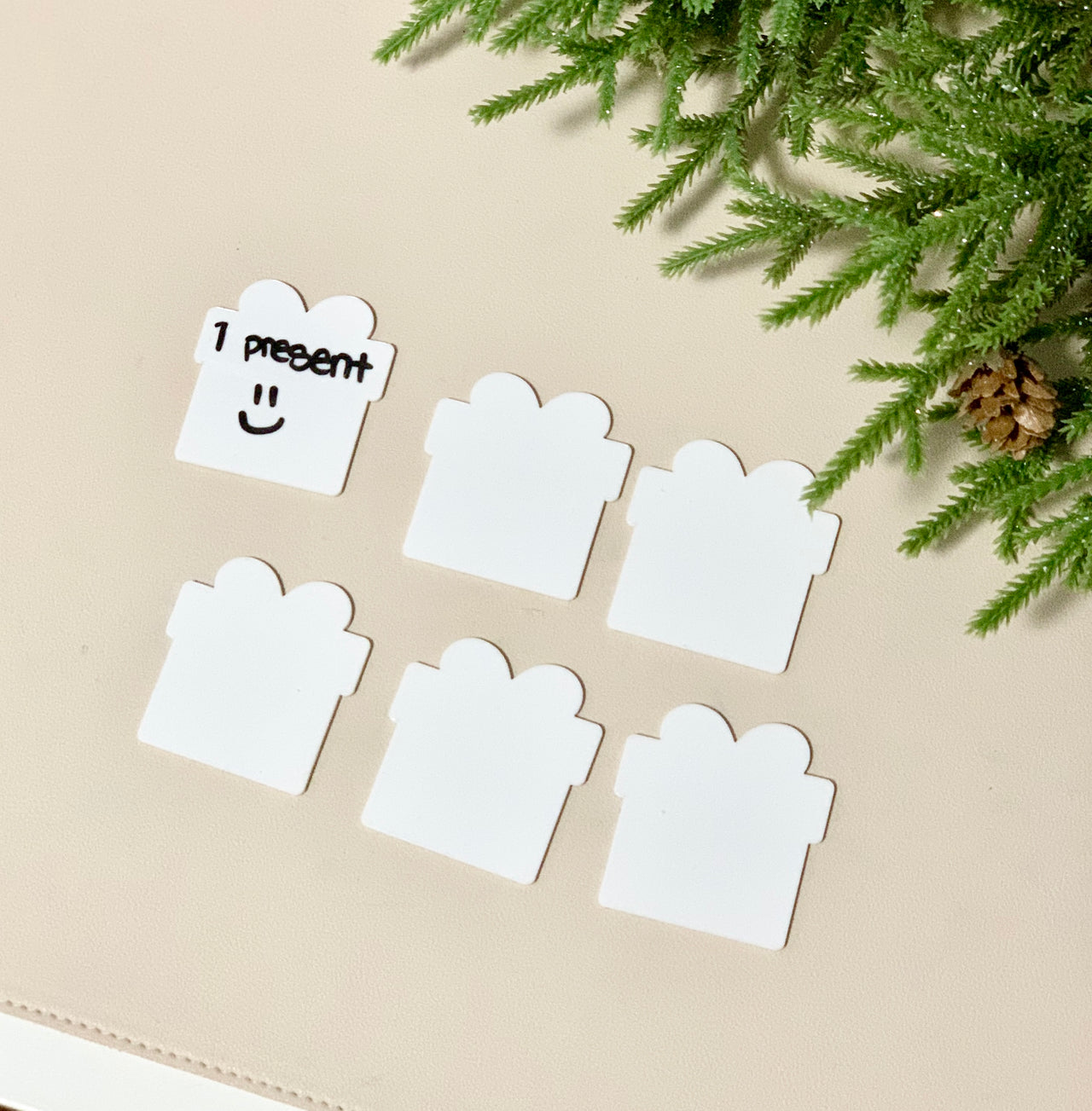 Set of six white little dry erasable cards shaped like presents on beige surface with pine branch decoration