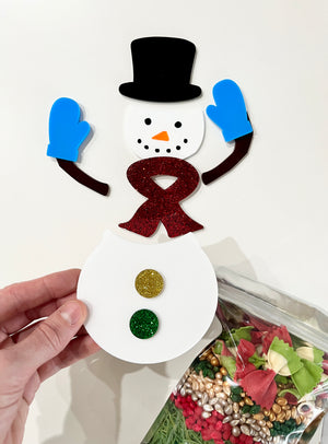 Hand holding white snowman craft piece with black hat, blue mittens, red glittery scarf, and colorful glitter buttons above festive pasta bag