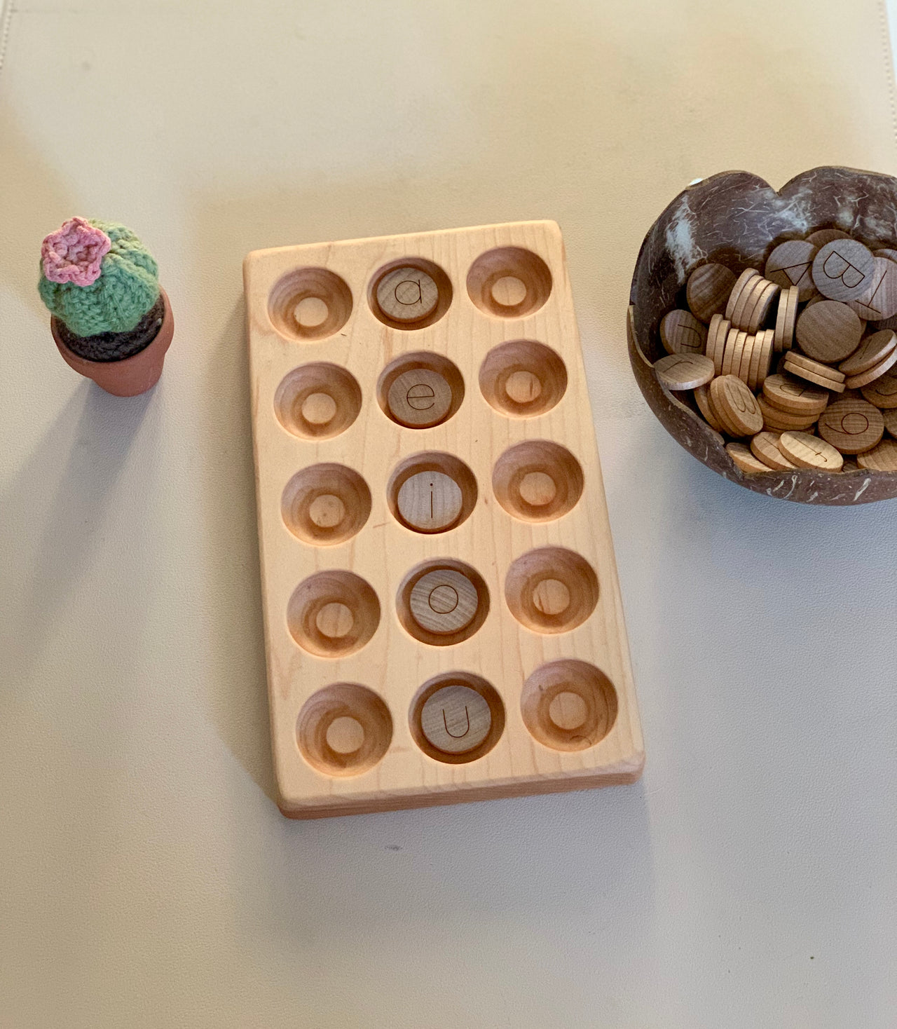 Light wooden tray with round indentations holding wooden letter pieces, next to a small crocheted cactus and coconut bowl with tokens