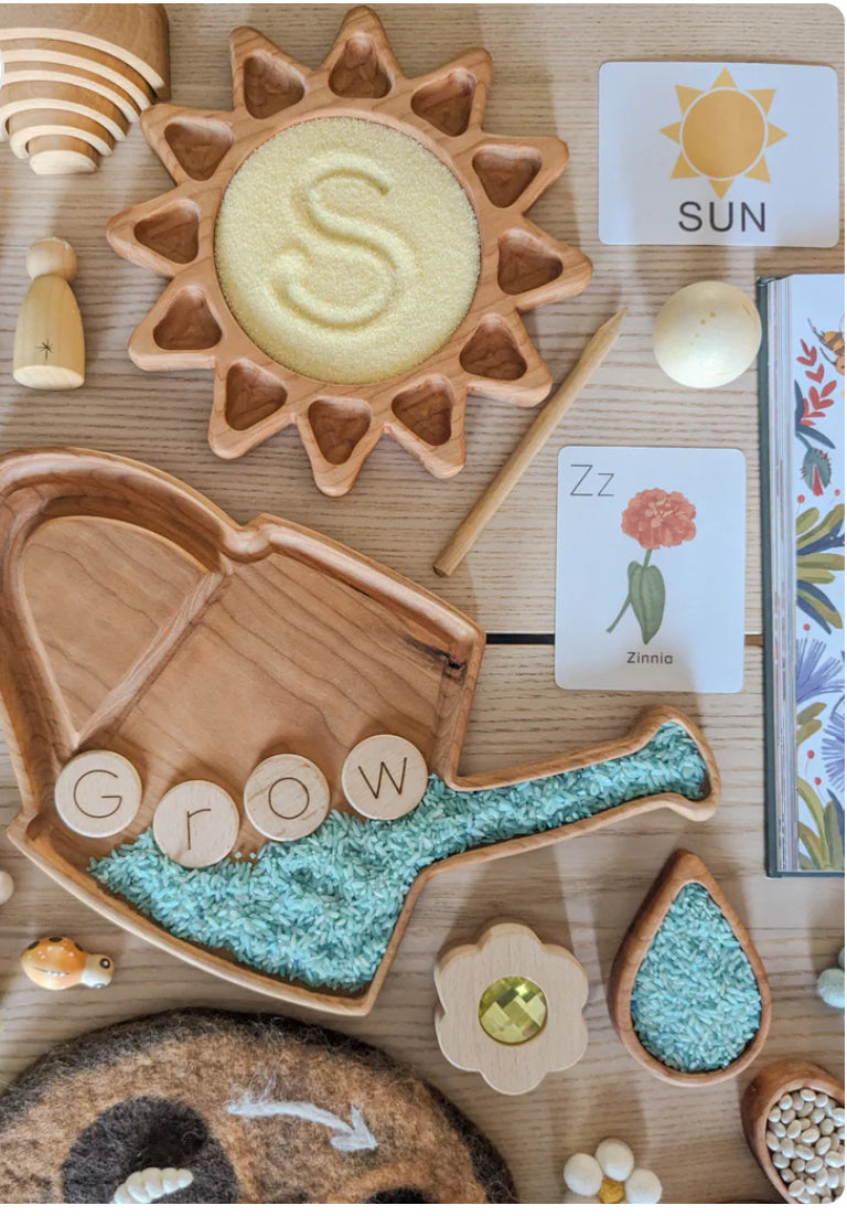 Wooden watering can sensory tray filled with blue rice and wooden letter circles spelling grow on light wood table