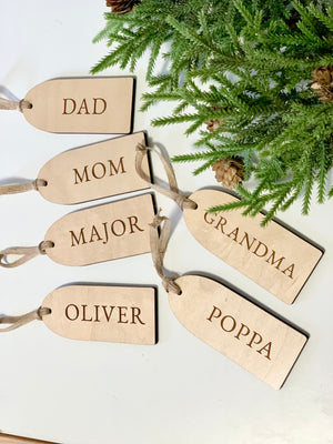 Set of six light wooden gift tags engraved with family names beside green pine branches and pinecones on white surface