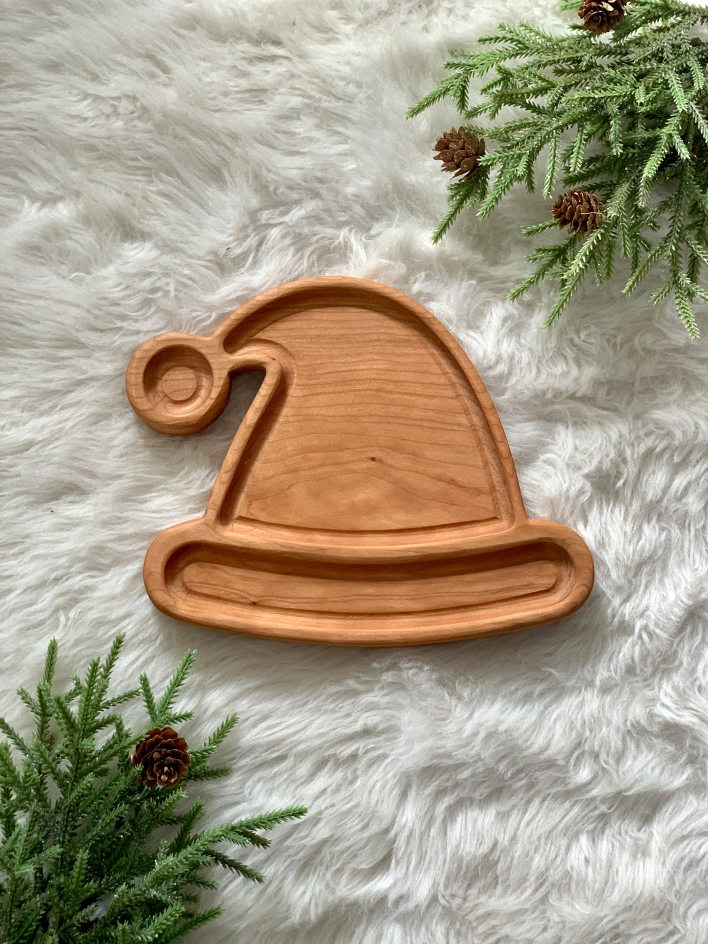 Wooden Santa or elf hat-shaped sensory tray on white faux fur with pine branches and pinecones nearby