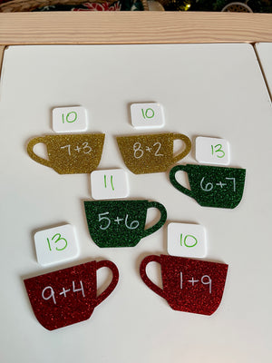 Glittered red, green, and gold coffee cup cutouts with addition problems and white tiles showing corresponding sums on a white surface