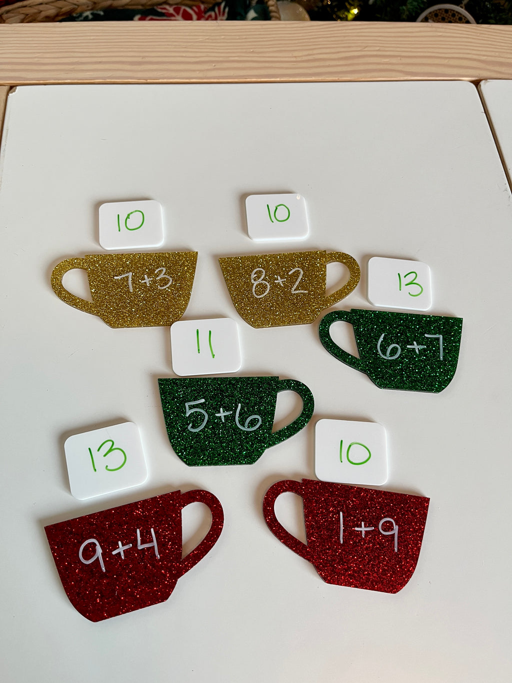 Glittered red, green, and gold coffee cup cutouts with addition problems and white tiles showing corresponding sums on a white surface