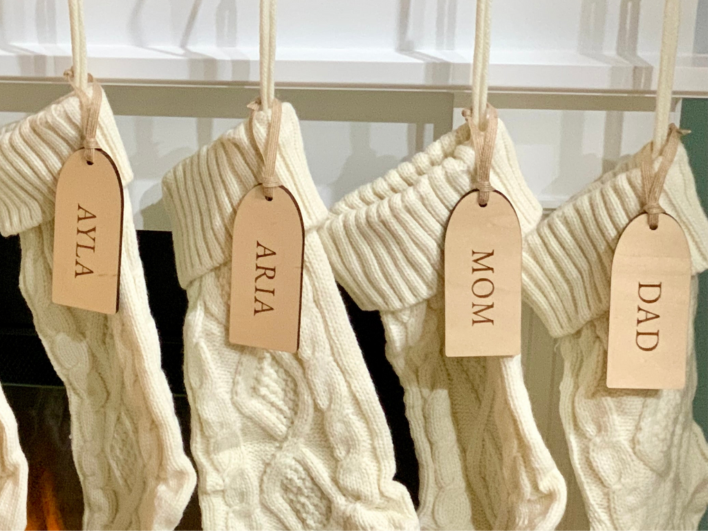 Four cream-colored knitted Christmas stockings hanging on a white mantle with leather name tags attached