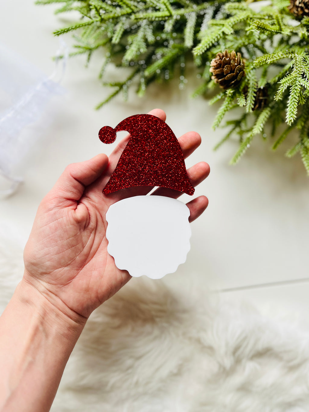 Hand holding red glitter Santa hat and white beard cutout with pinecone and greenery background on light surface