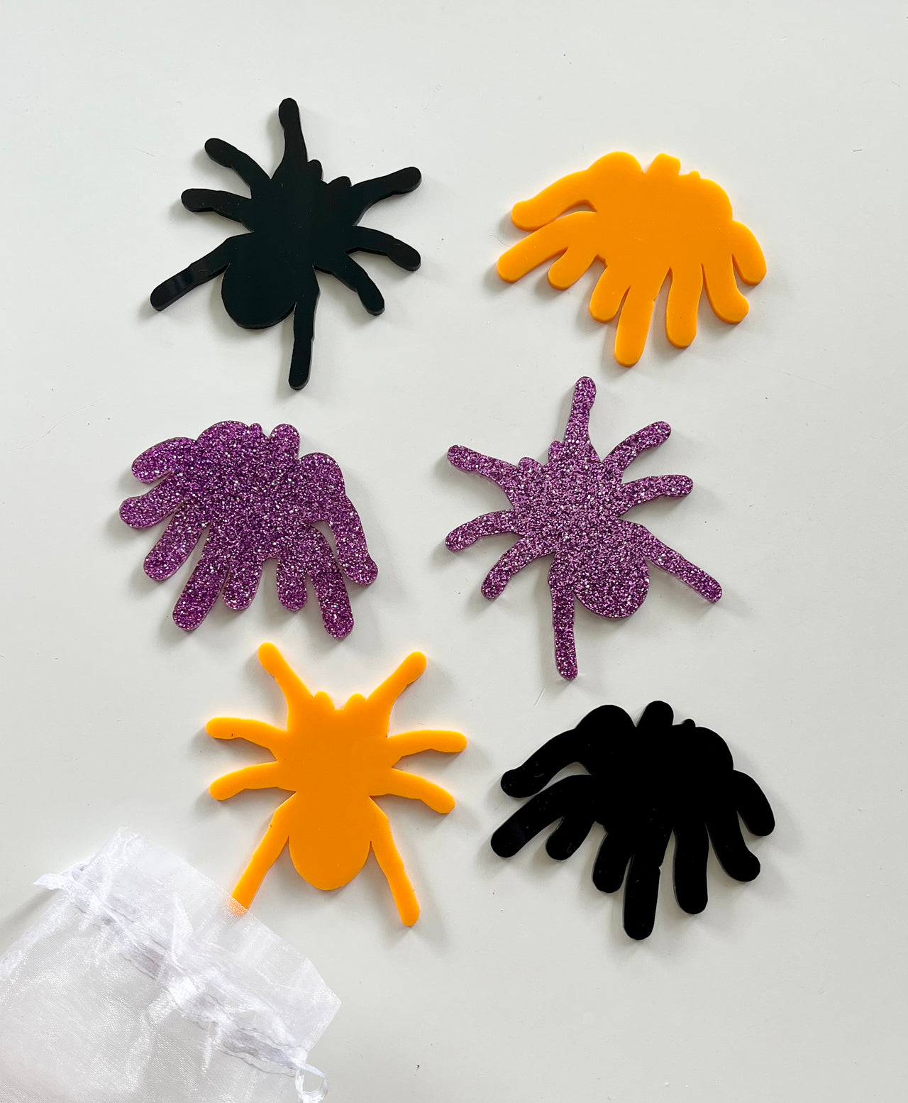 Six spider-shaped cutouts in black, orange, and purple glitter arranged on a white surface near a sheer white bag