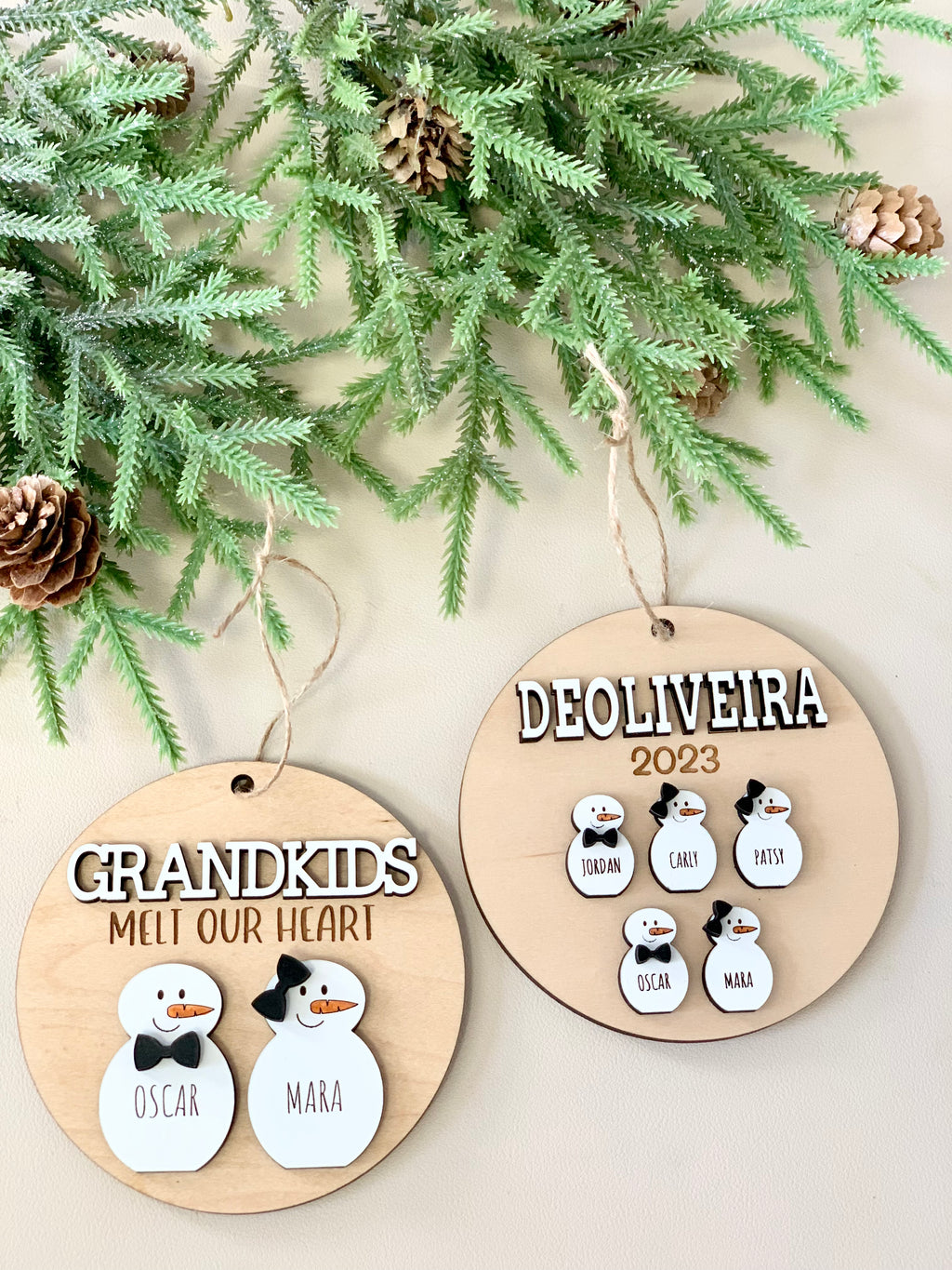 Personalized wooden Christmas ornaments with snow person figures and names, hanging on a pine branch background