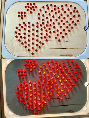 Wooden board with four heart outlines filled with removable red pins, shown unlit and glowing in a light box.