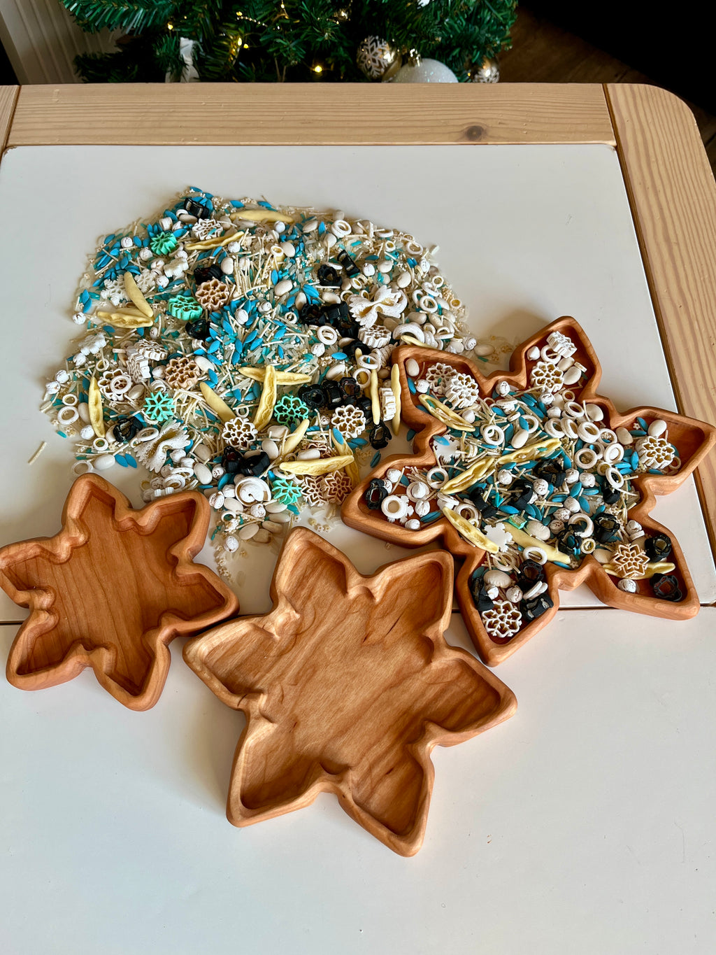 Wooden leaf-shaped trays with assorted winter-themed sensory materials including seeds, beads, and dried botanicals on white table