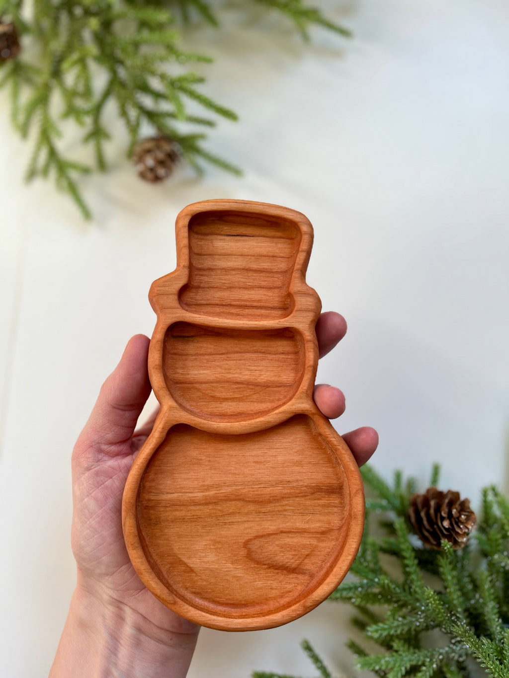 Hand holding a carved wooden tray shaped like a snowman with pine branches and pine cones nearby on white surface