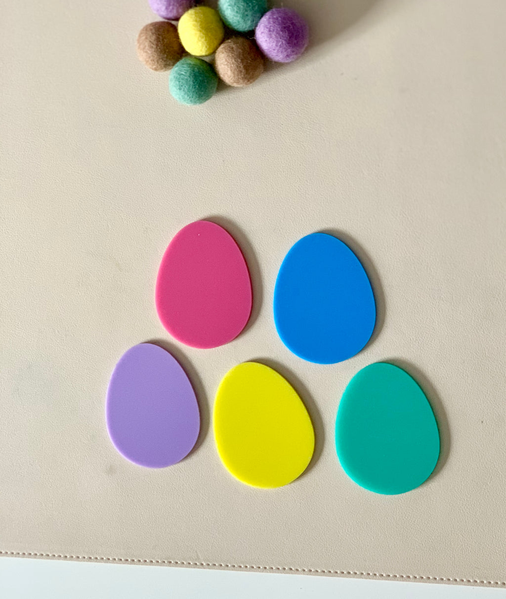 Five colorful egg-shaped pieces arranged in a semicircle on a light surface with felt balls above them