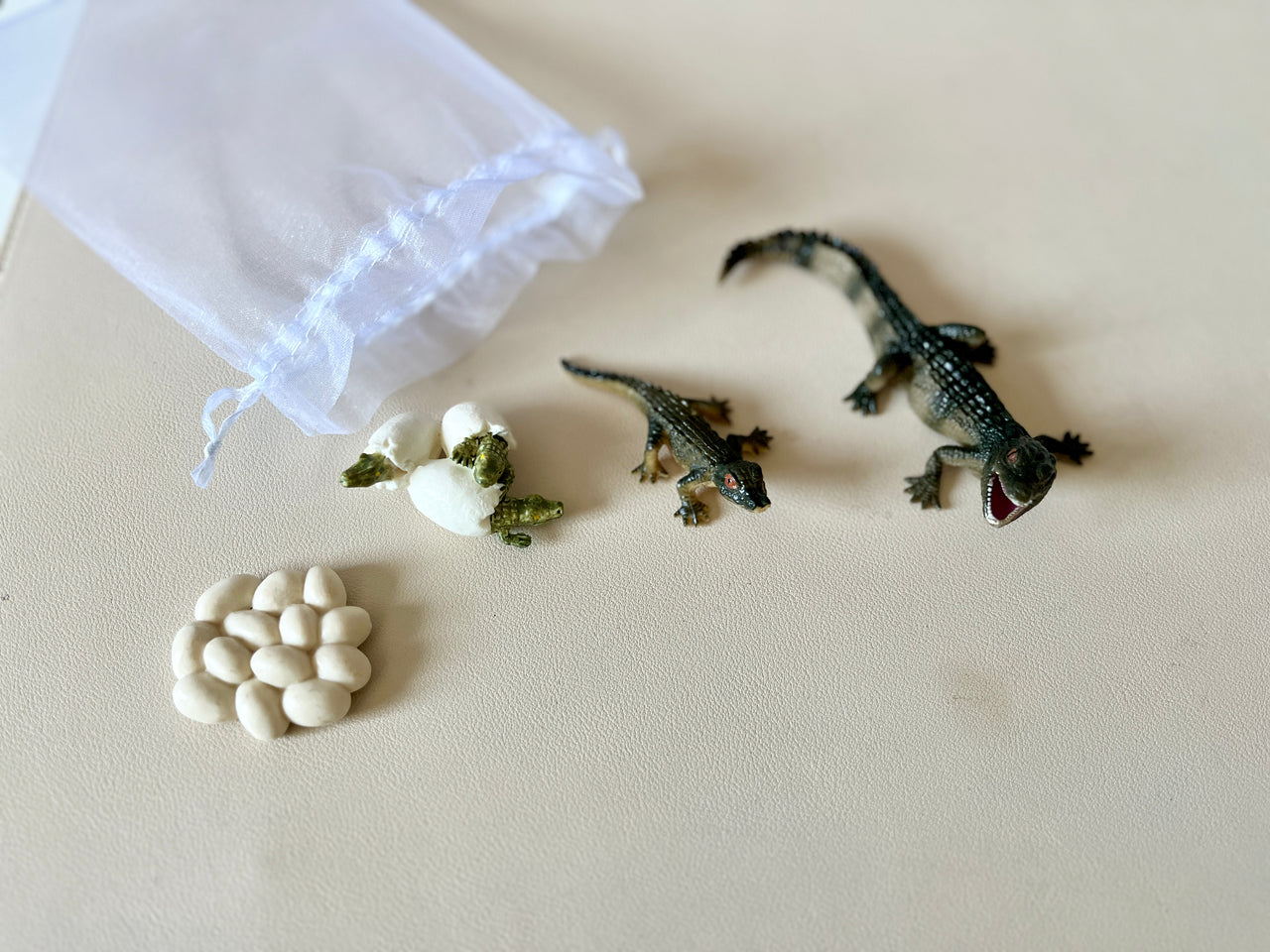 Plastic alligator figurines, white egg models, and seed replicas near a white sheer drawstring bag on beige surface