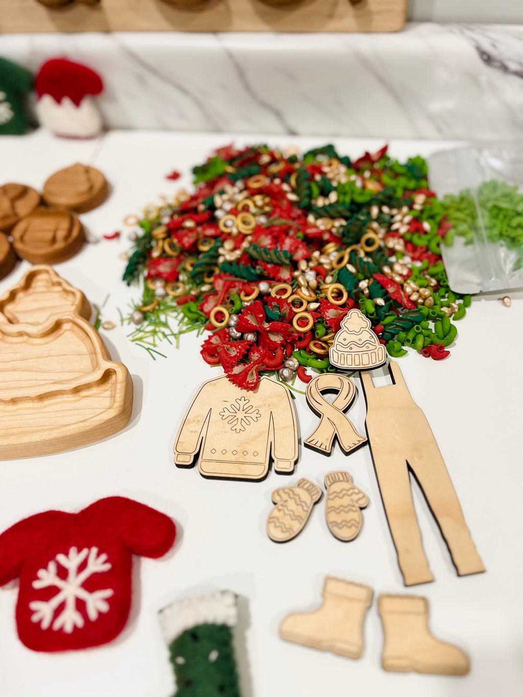 Wooden cutouts of winter clothing including sweater, mittens, boots, and hat next to colorful spiral pasta on white surface