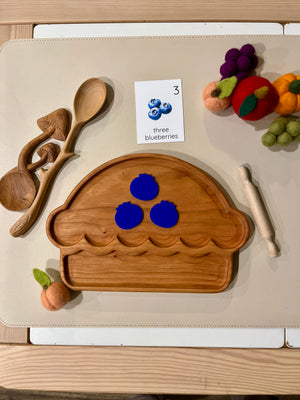 Wooden pie-shaped tray with blue pomegranate cutouts, wooden spoons, mini rolling pin, and felt fruit on beige surface