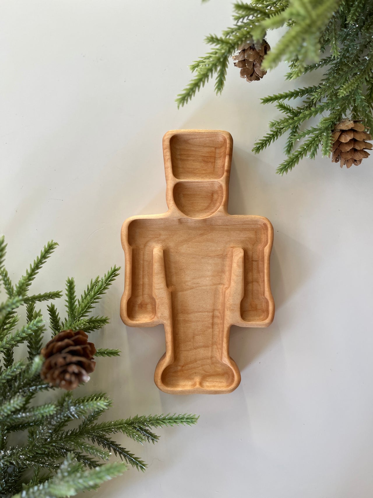 Light wooden tray carved in a robot shape surrounded by pine branches and pinecones on a white background