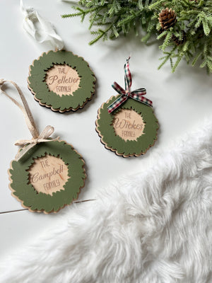 Three layered wooden wreath ornaments with green leaves and personalized family names on white surface next to greenery and fur rug