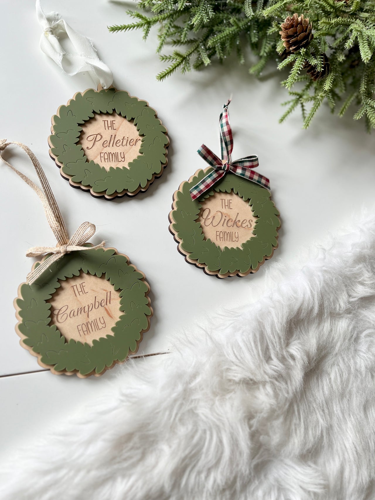 Three layered wooden wreath ornaments with green leaves and personalized family names on white surface next to greenery and fur rug