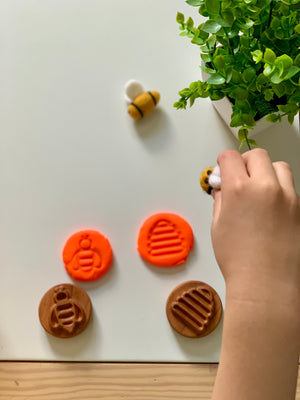 Child’s hand holding felted bee toy near wooden stamps and matching orange clay impressions on white surface with plant nearby