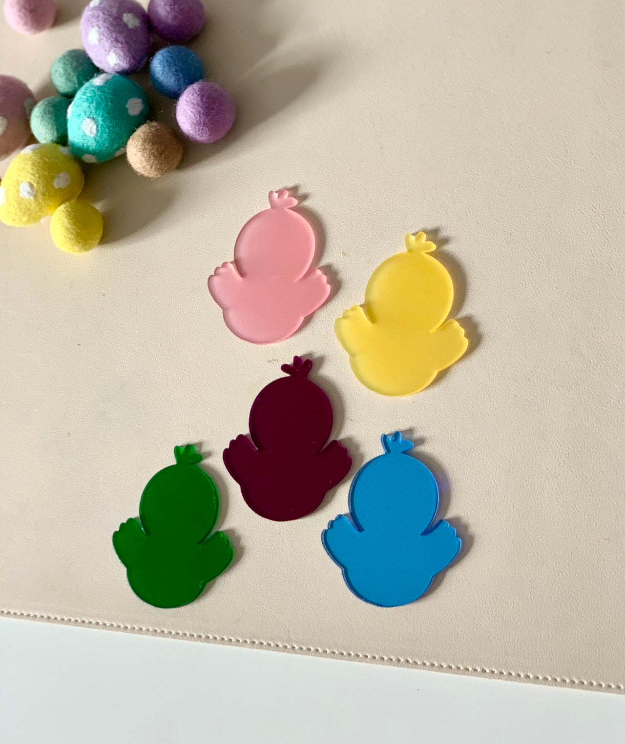 Five translucent bird-shaped cutouts in pink, yellow, burgundy, green, and blue on a beige surface near felt balls