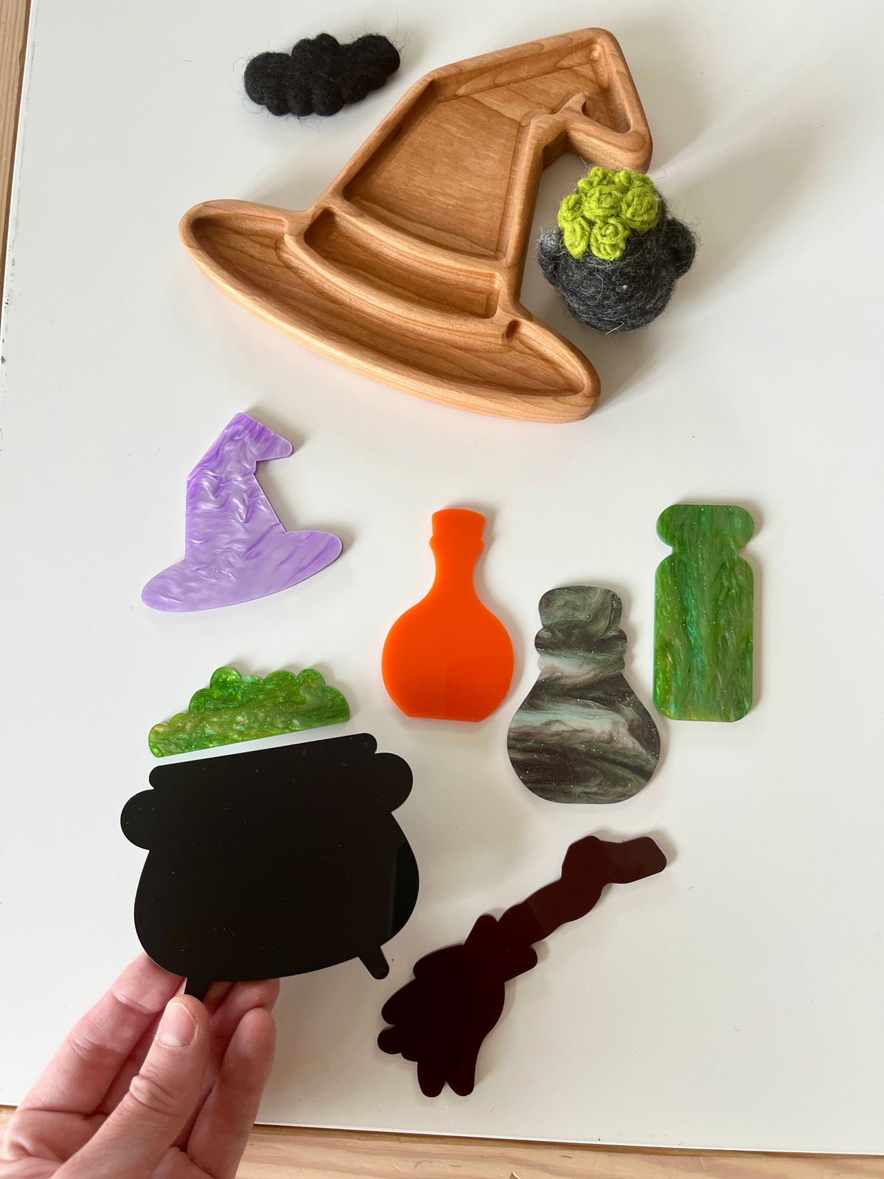Wooden witch hat tray with assorted colorful acrylic potion bottles, cauldron shape, broom, and felt decorations on white surface