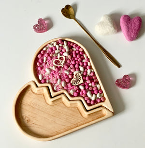 Broken heart-shaped wooden tray filled with pink and white sensory beads, felt hearts, and a gold heart-topped spoon nearby