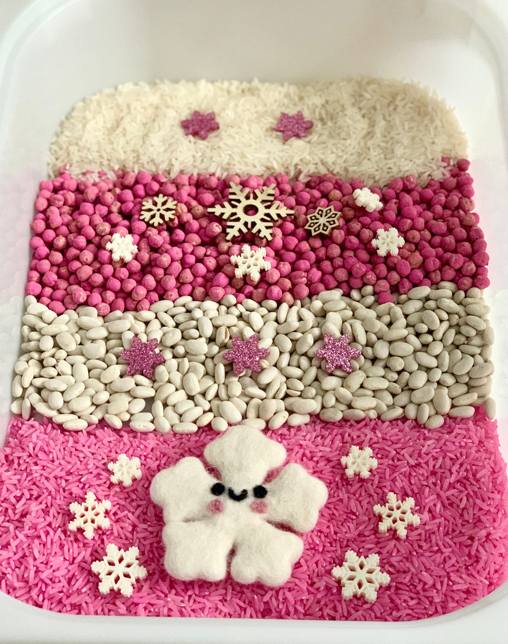 Layered sensory bin with pink rice, white beans, pink beads, wooden and felt snowflakes, and a smiling felt snowflake figure