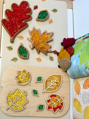 Wooden leaf-shaped sensory trays filled with colorful beads, felt leaves, and an open book on white surface