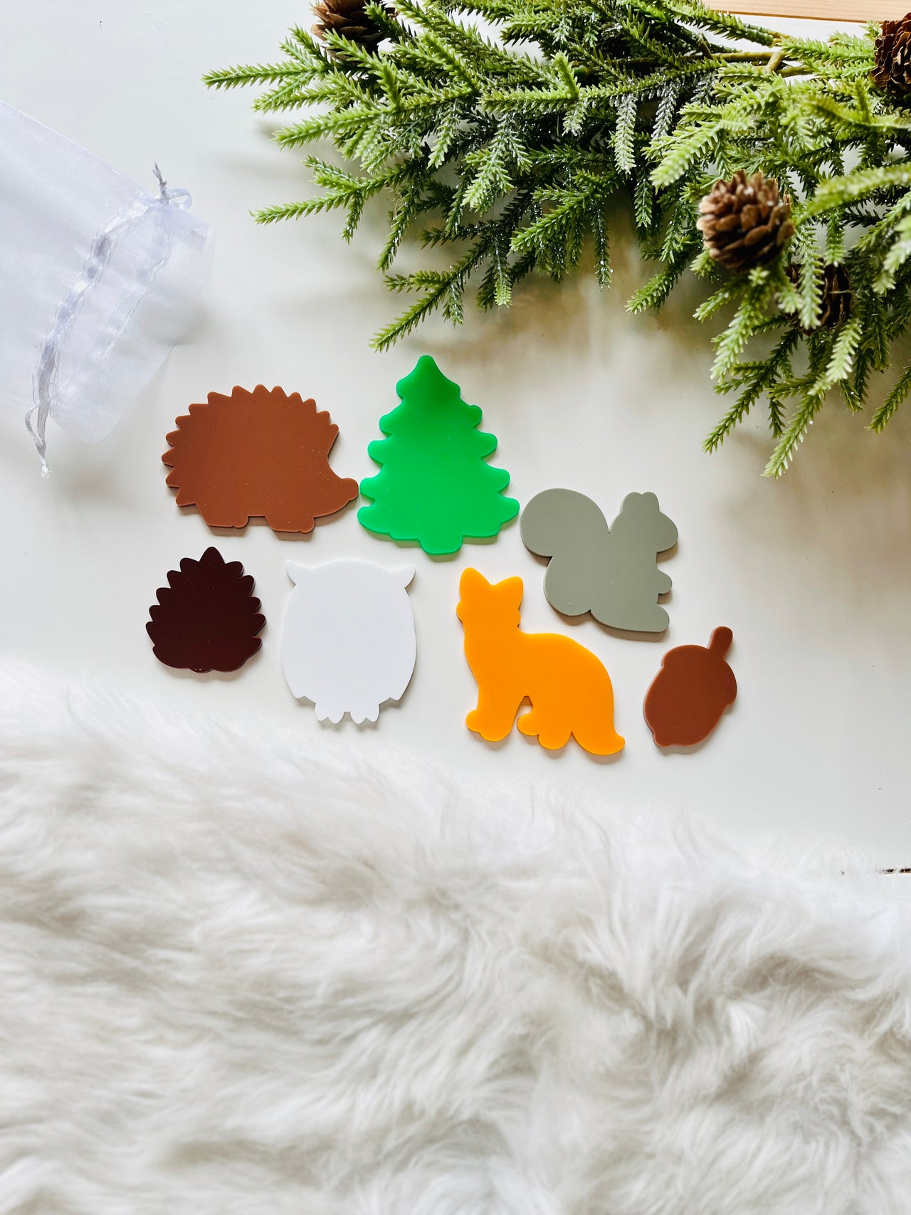Colorful flat acrylic shapes of a hedgehog, tree, squirrel, pinecone, owl, fox, and acorn near pine branches and white fur background