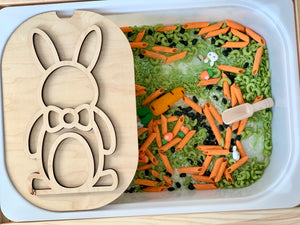 Plastic bin filled with green and orange pasta shapes and a wooden bunny stencil resting on the edge