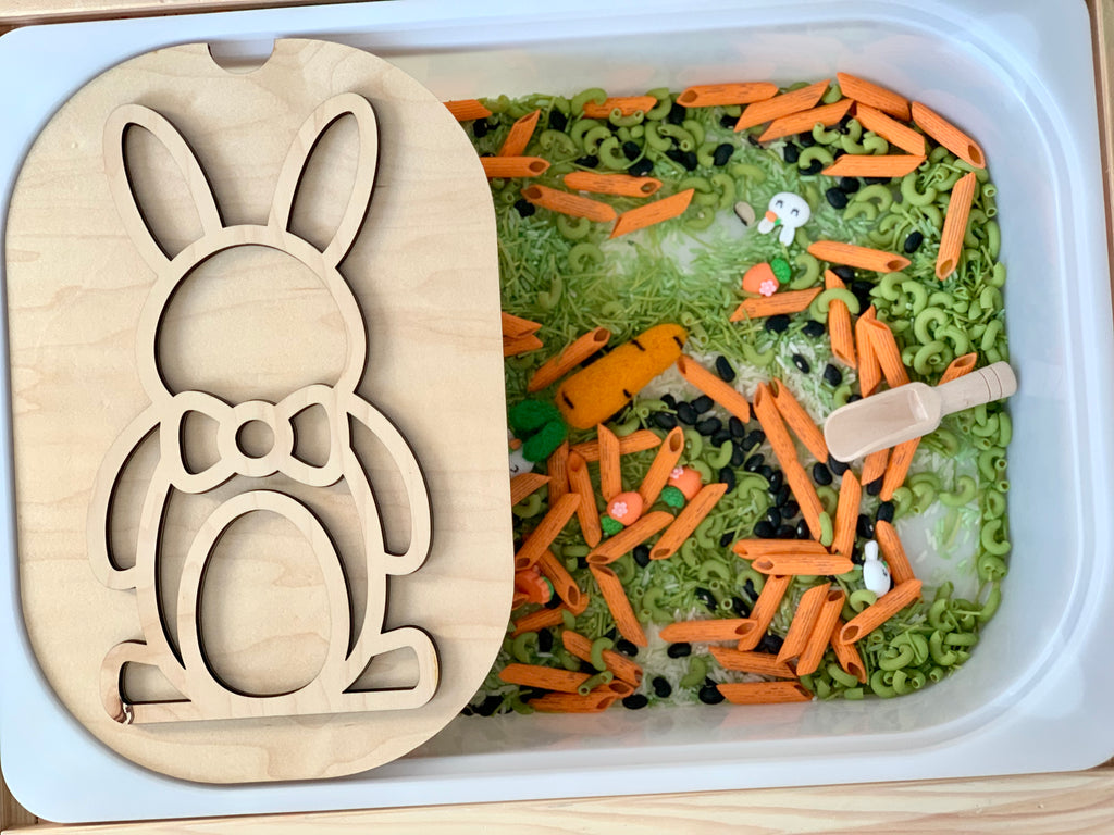 Plastic bin filled with green and orange pasta shapes and a wooden bunny stencil resting on the edge