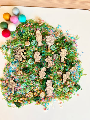 Wooden Nutcracker and ballerina cutouts on green pine needle mix with colorful sprinkles and small gold beads on white surface