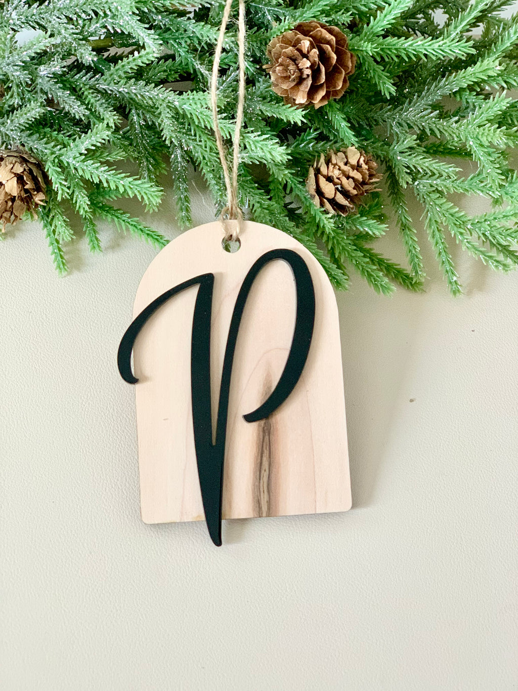 Light wood arch-shaped ornament with a black cursive letter P hanging from twine against pine branches and pinecones