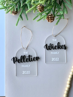 Two clear acrylic arch ornaments with black cursive family names and white text showing years, hanging from twine below pine branches