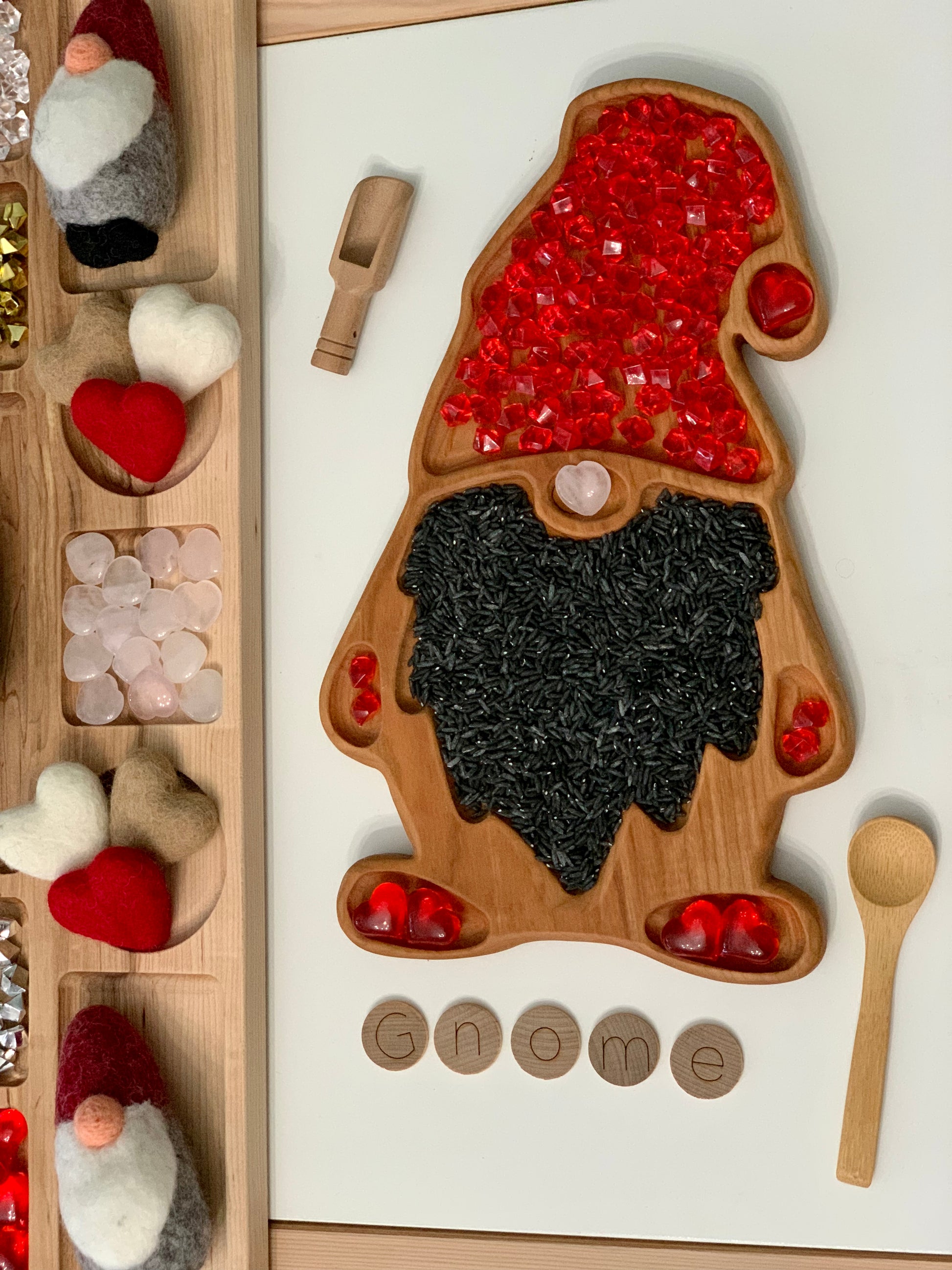 Wooden gnome-shaped sensory tray filled with red gems and black rice with wooden scoop and small wooden spoon nearby