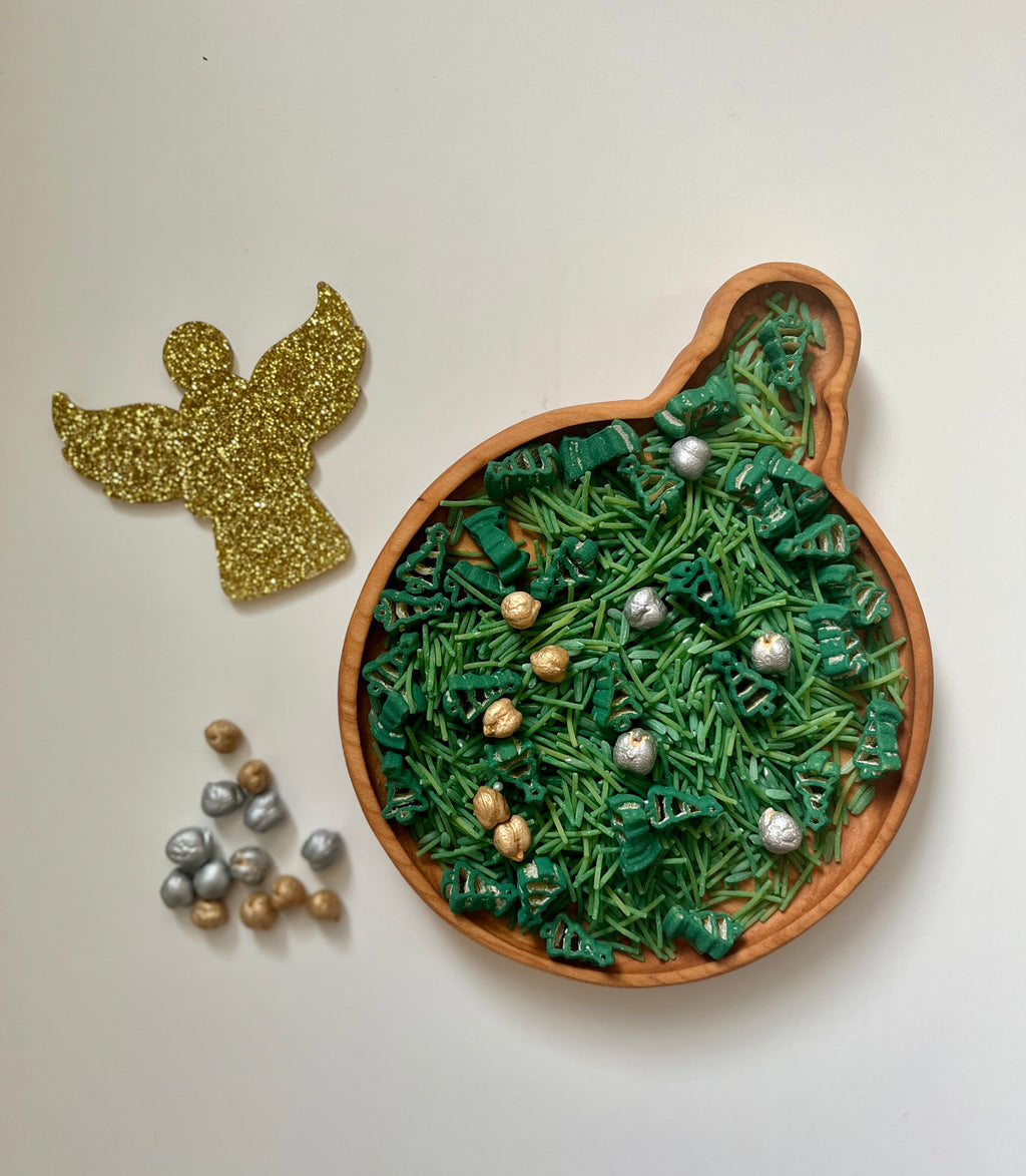 Round wooden tray shaped like a Christmas ornament filled with green shredded paper and small silver and gold decorative balls next to a glittery gold angel cutout