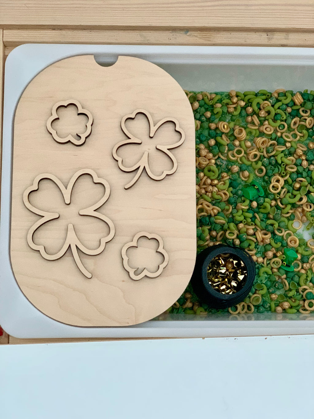 Wooden board with shamrock cutouts beside tray of green and beige pasta, small green shapes, and a black pot with gold pieces